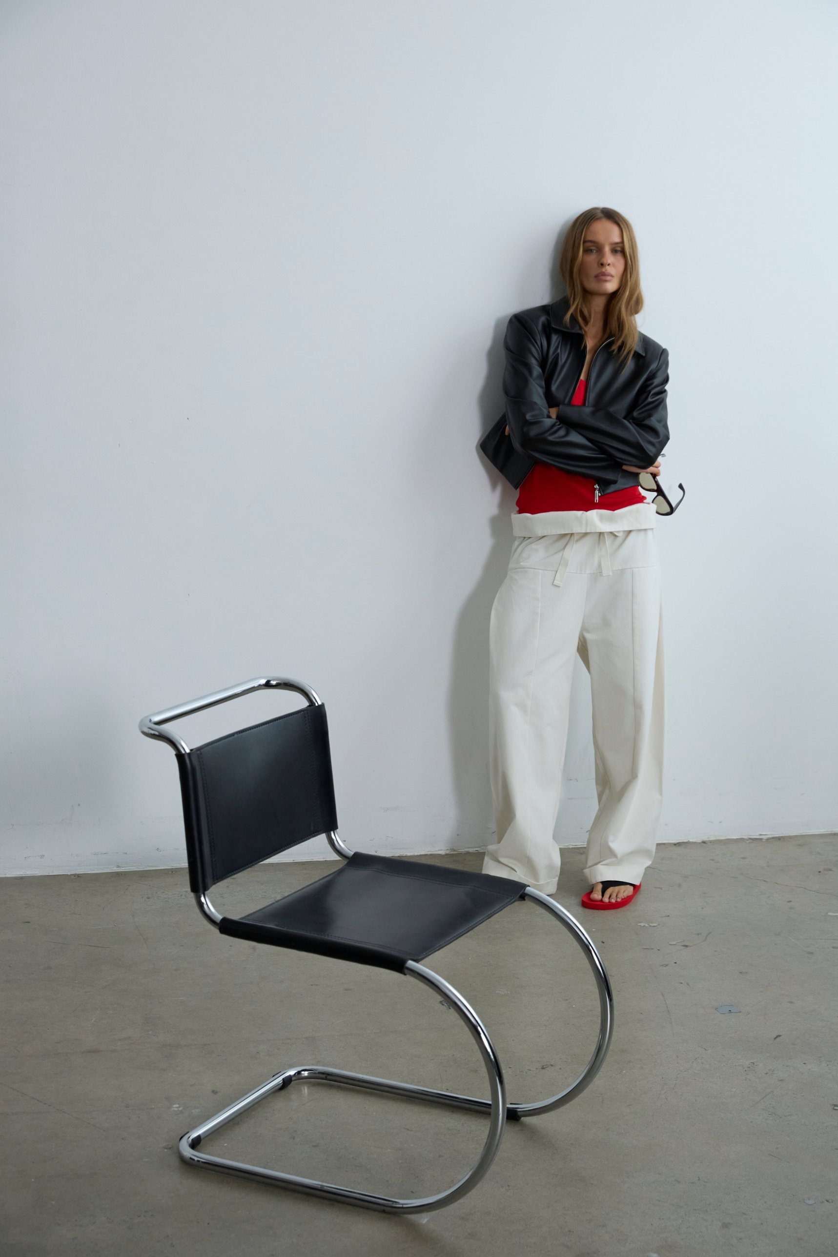 A woman in a black leather jacket, red top and cream drawstring trousers, leaning against a wall.